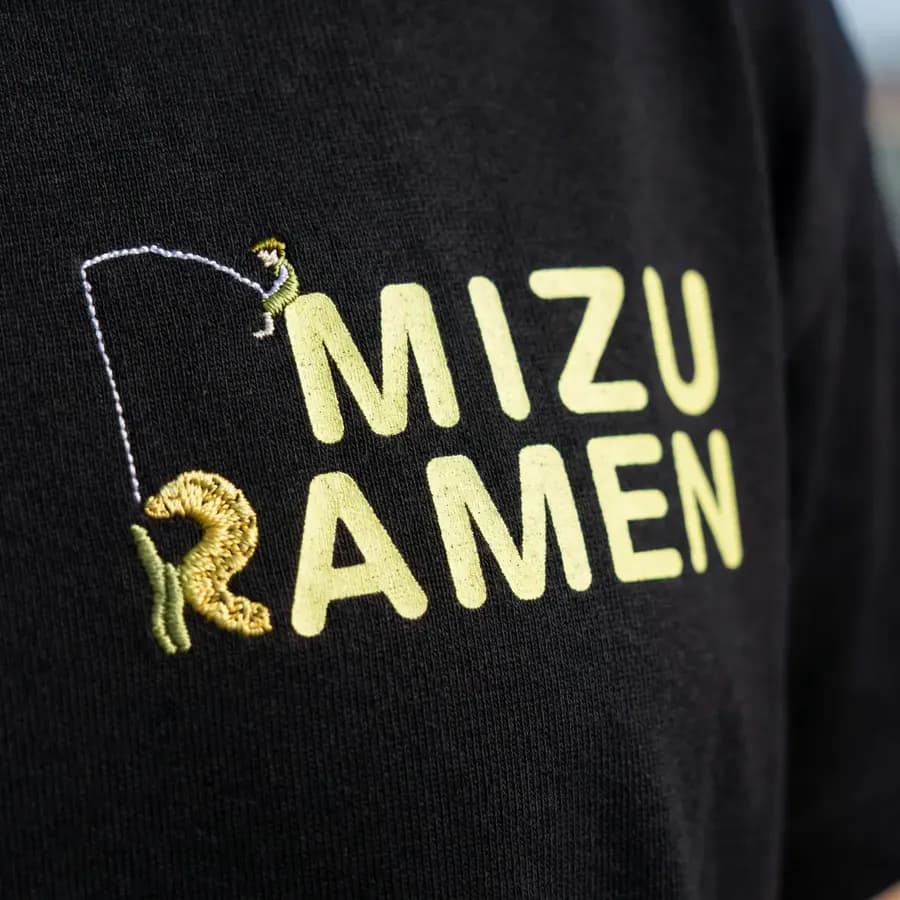 黒いTシャツの胸ロゴの接写。文字はプリント、釣り人と「R」の部分は刺繍